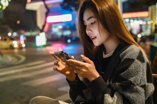 Free A young woman enjoys her smartphone amidst the lively ambiance of a city street at night. Stock Photo