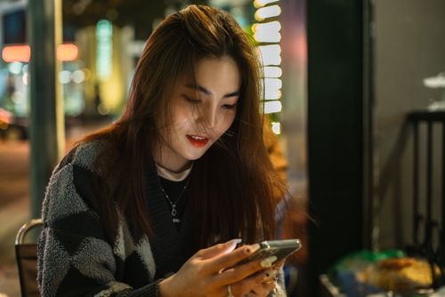 Free A young woman with brunette hair sits outdoors at night, texting on her phone in an urban setting. Stock Photo