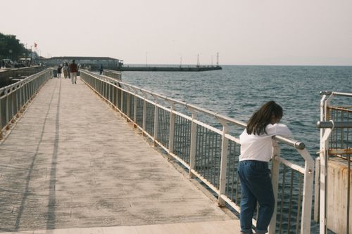 Free A child stands thoughtfully on a pier, looking over the calm ocean on a sunny day. Stock Photo