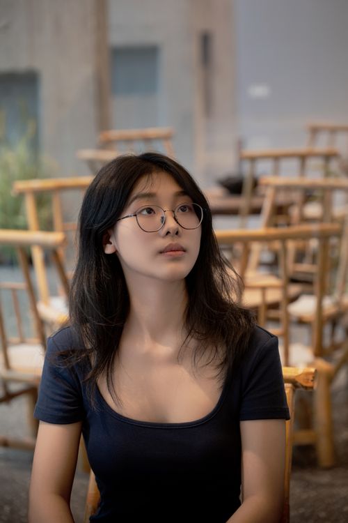 Free A thoughtful young woman with eyeglasses sits indoors, surrounded by wooden chairs. Stock Photo