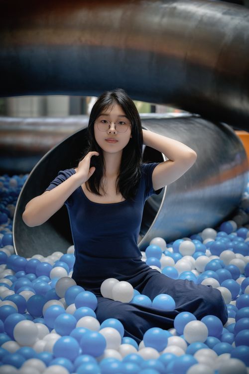 Free A fashionable woman with glasses sits in a ball pit, surrounded by colorful balls. Stock Photo