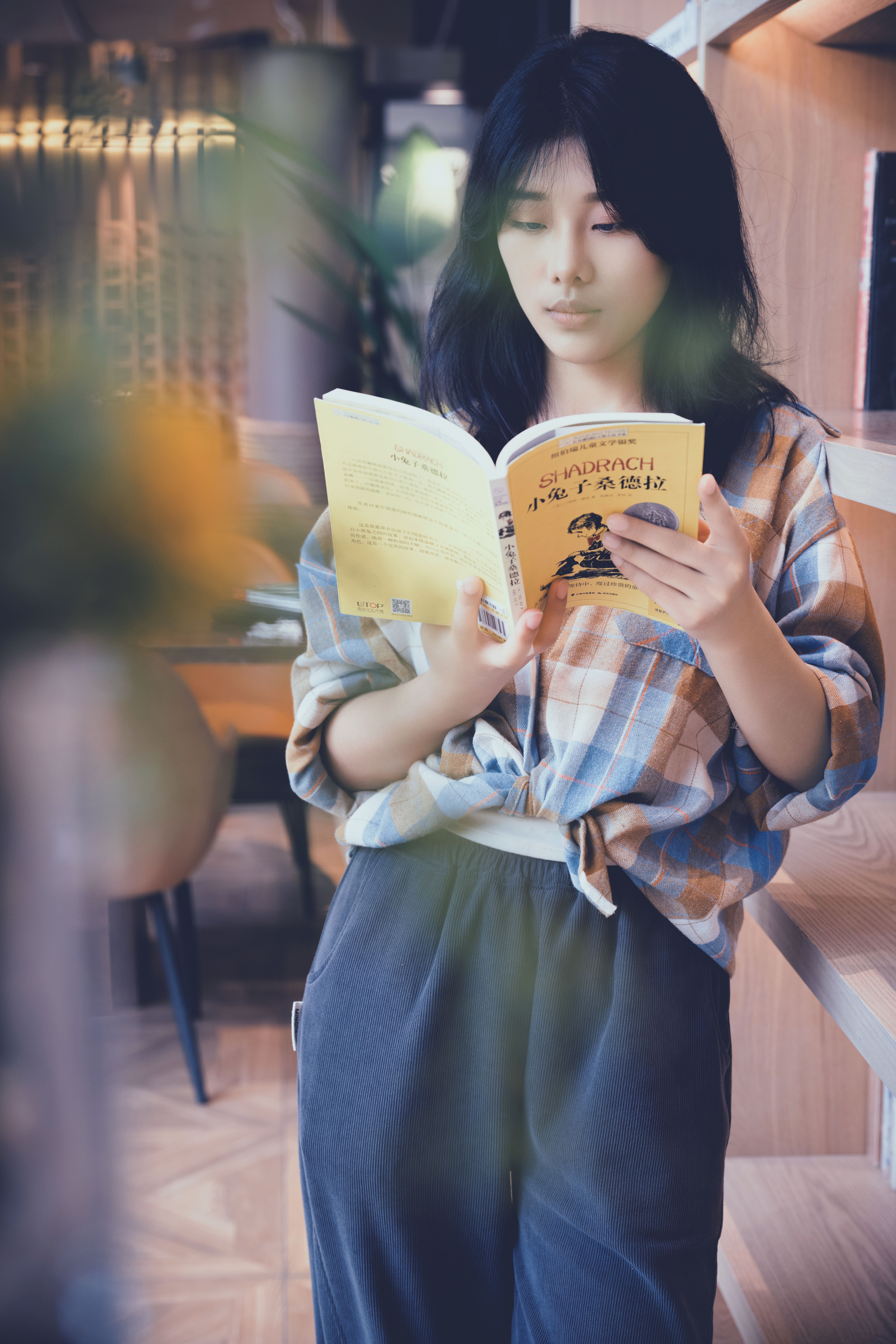 Asian woman reading a book indoors, wearing a plaid shirt. Relaxed atmosphere.