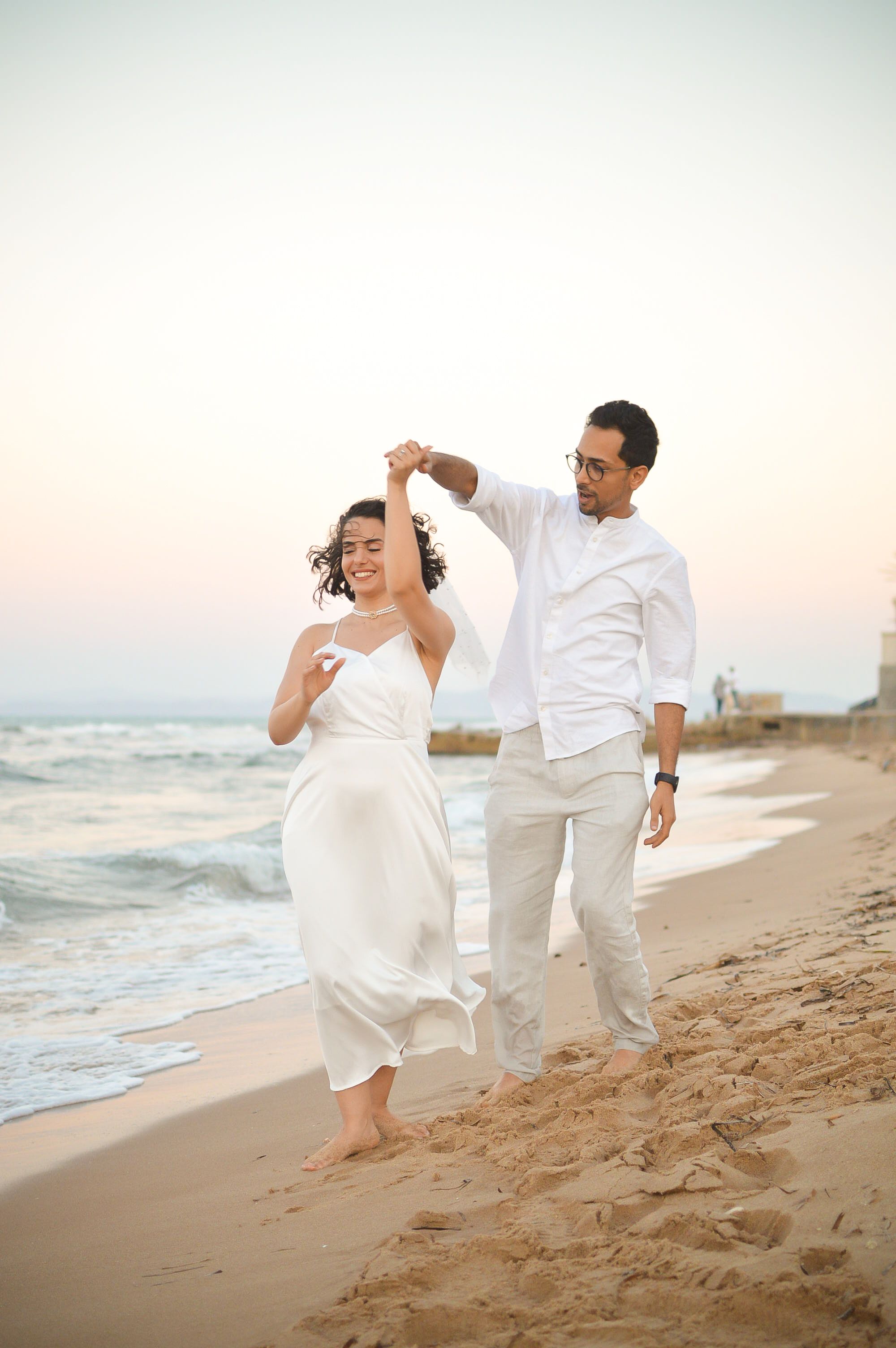 A joyful couple in white attire walking by the ocean shore, embodying love and happiness.