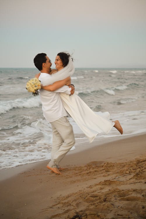 A bride and groom are hugging on the beach