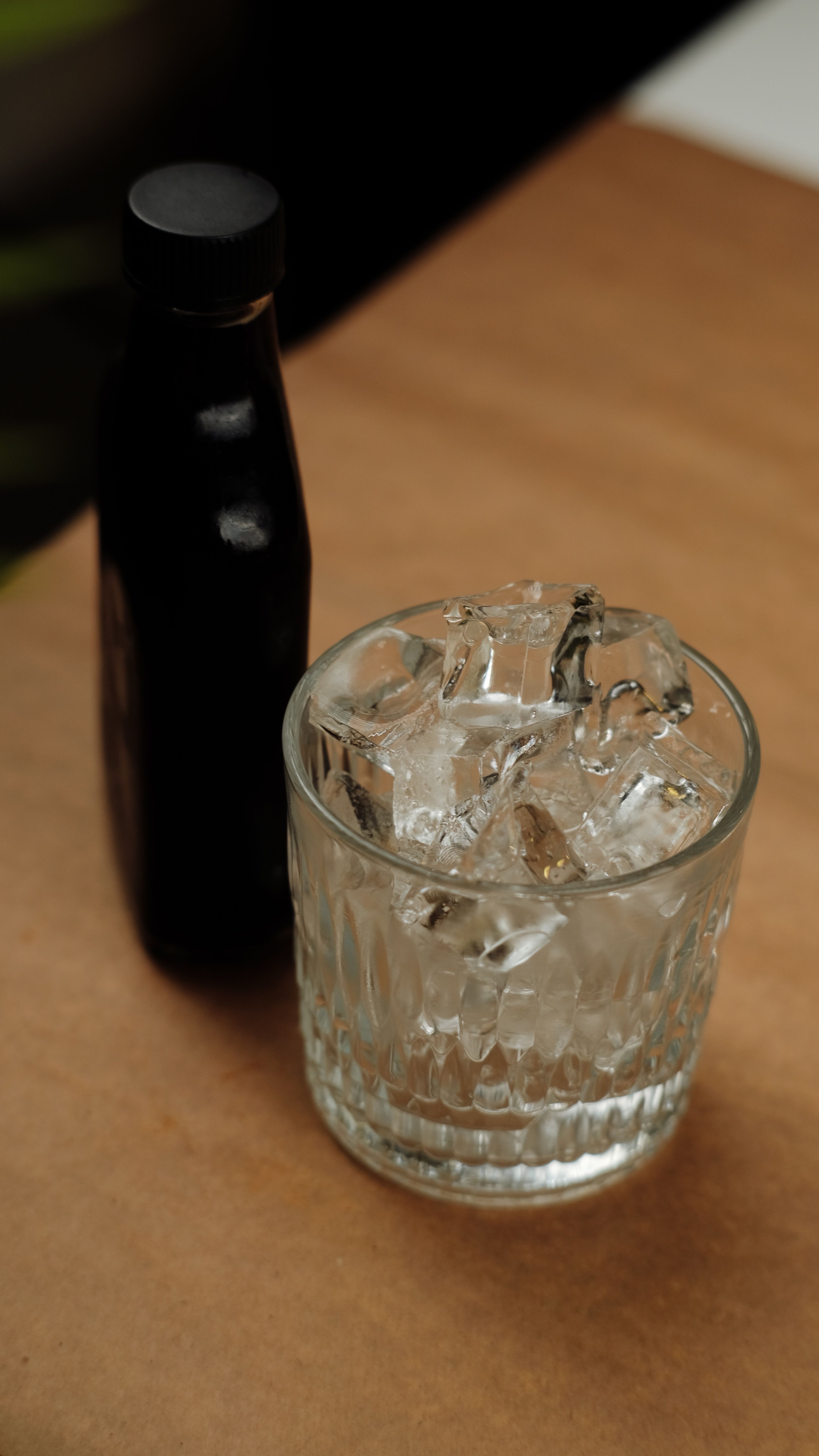 A close-up view of a glass filled with ice cubes and a black bottle on a wooden surface.