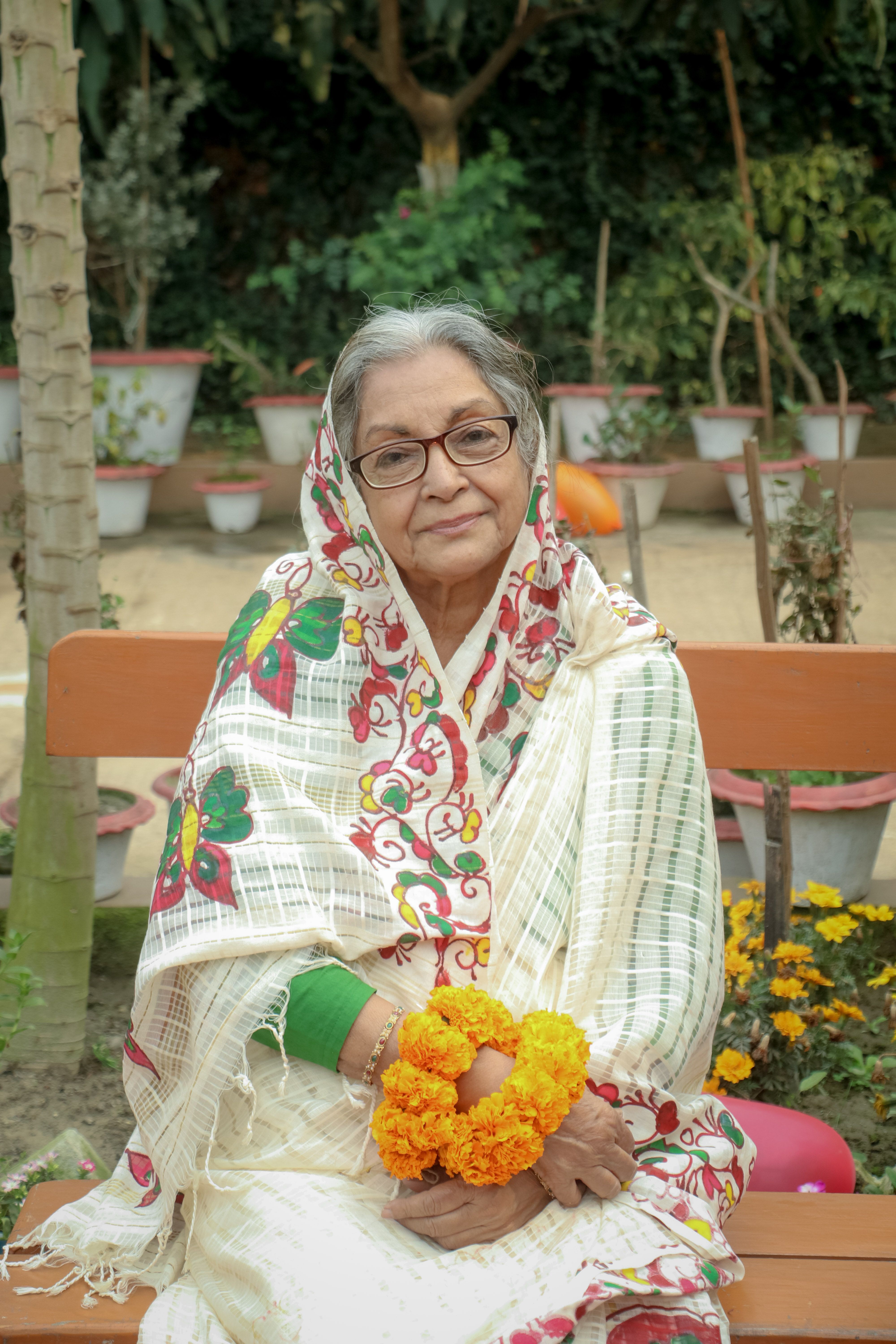 Elderly woman in Dhaka garden with flower bracelet, wearing traditional attire.