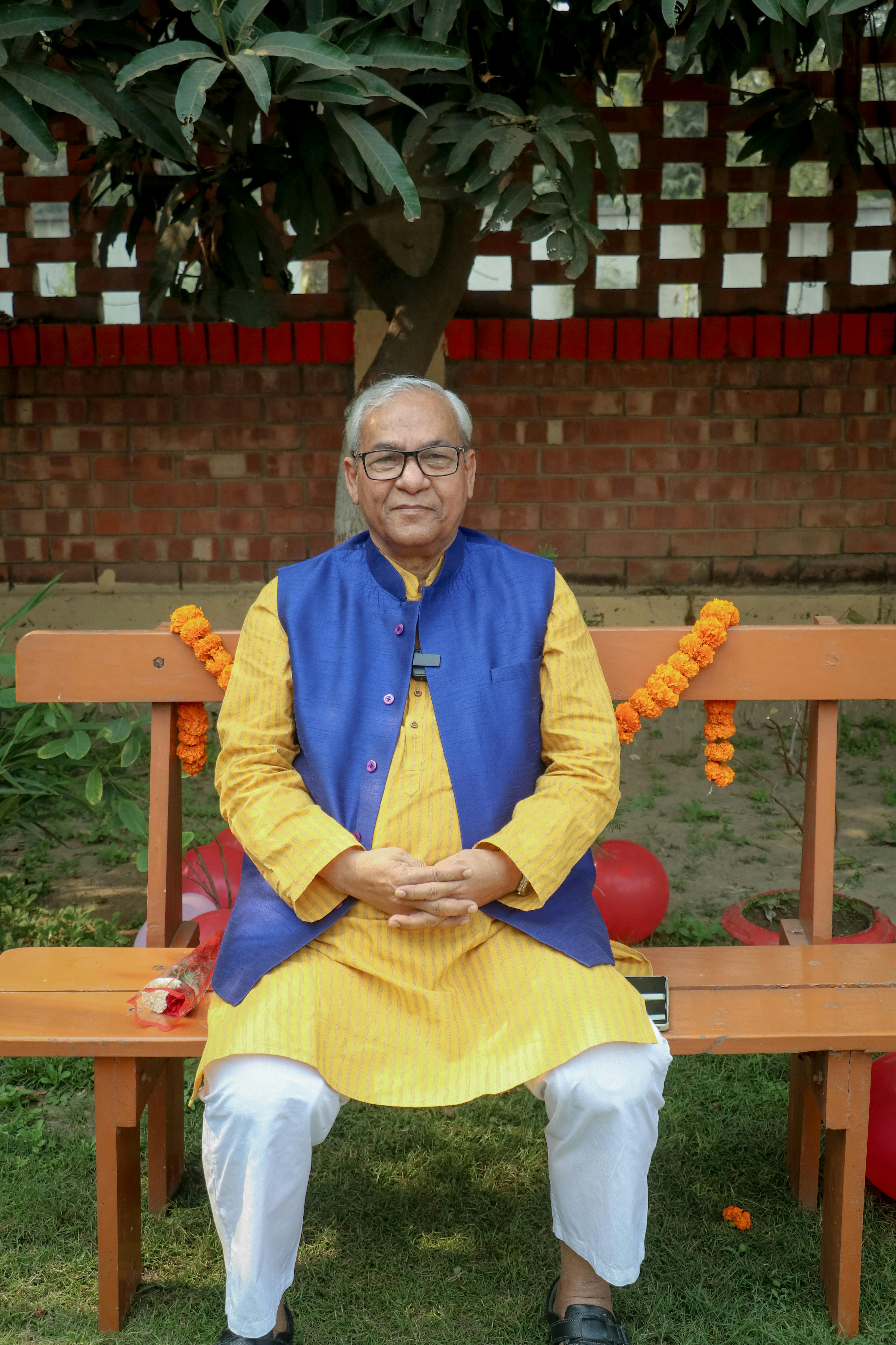 Senior Indian man in vibrant traditional attire sitting outdoors on a park bench.