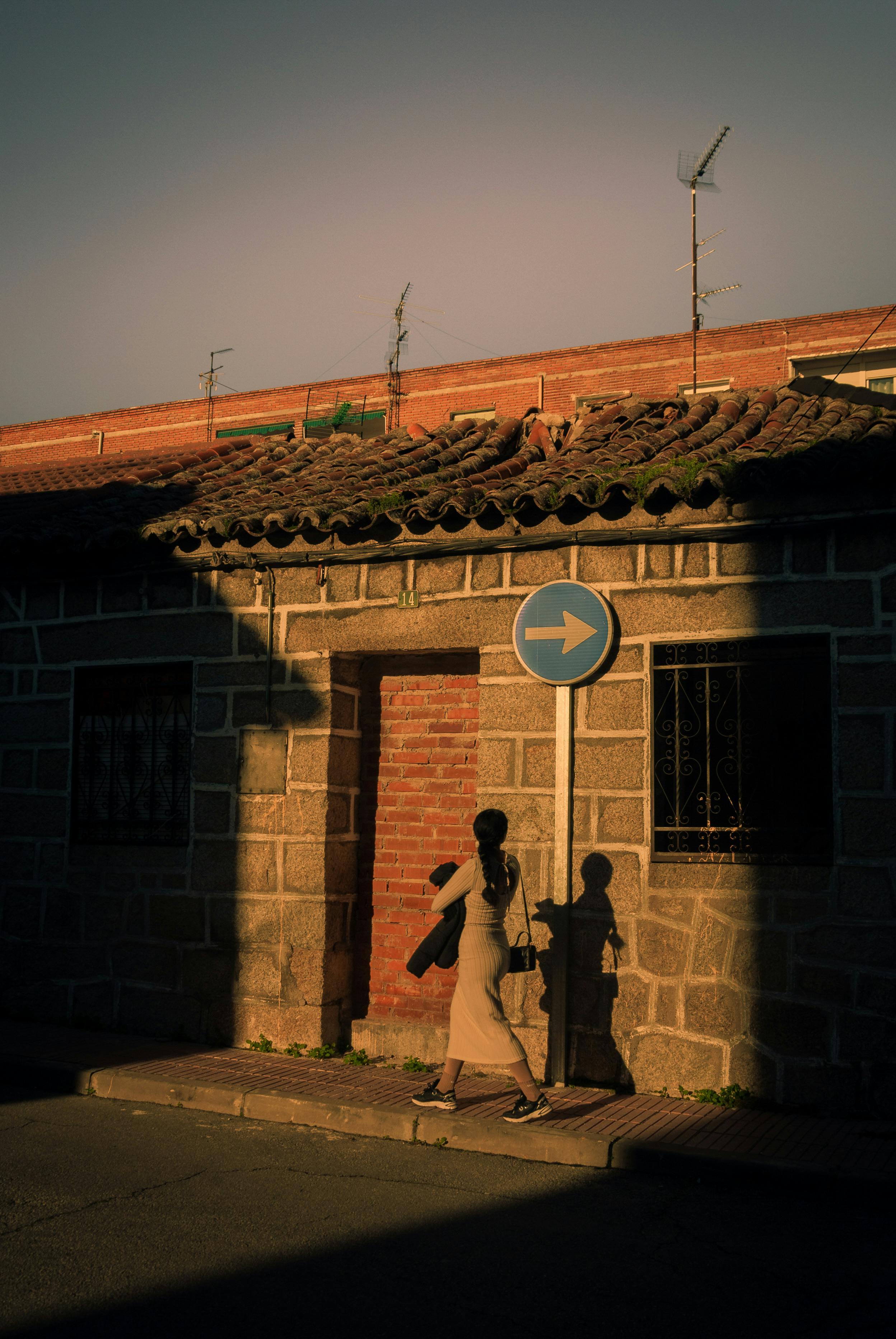 A woman walks past a building in a village at sunset, casting a shadow on the wall.