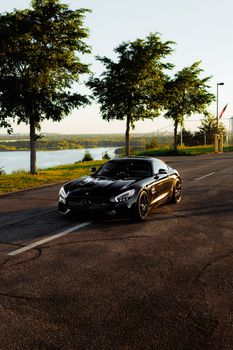 Black sports car parked on an empty road with trees and a lake view at sunrise.