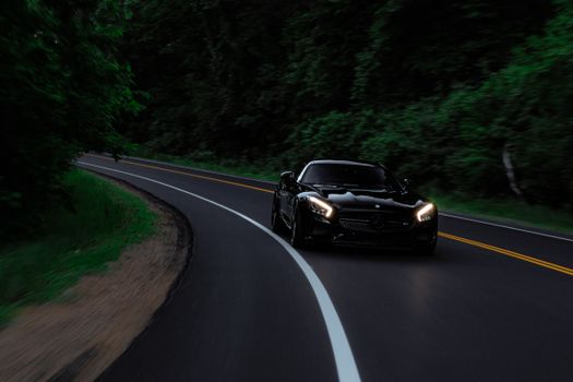 Black sports car driving through a winding forest road, headlights illuminating the path.