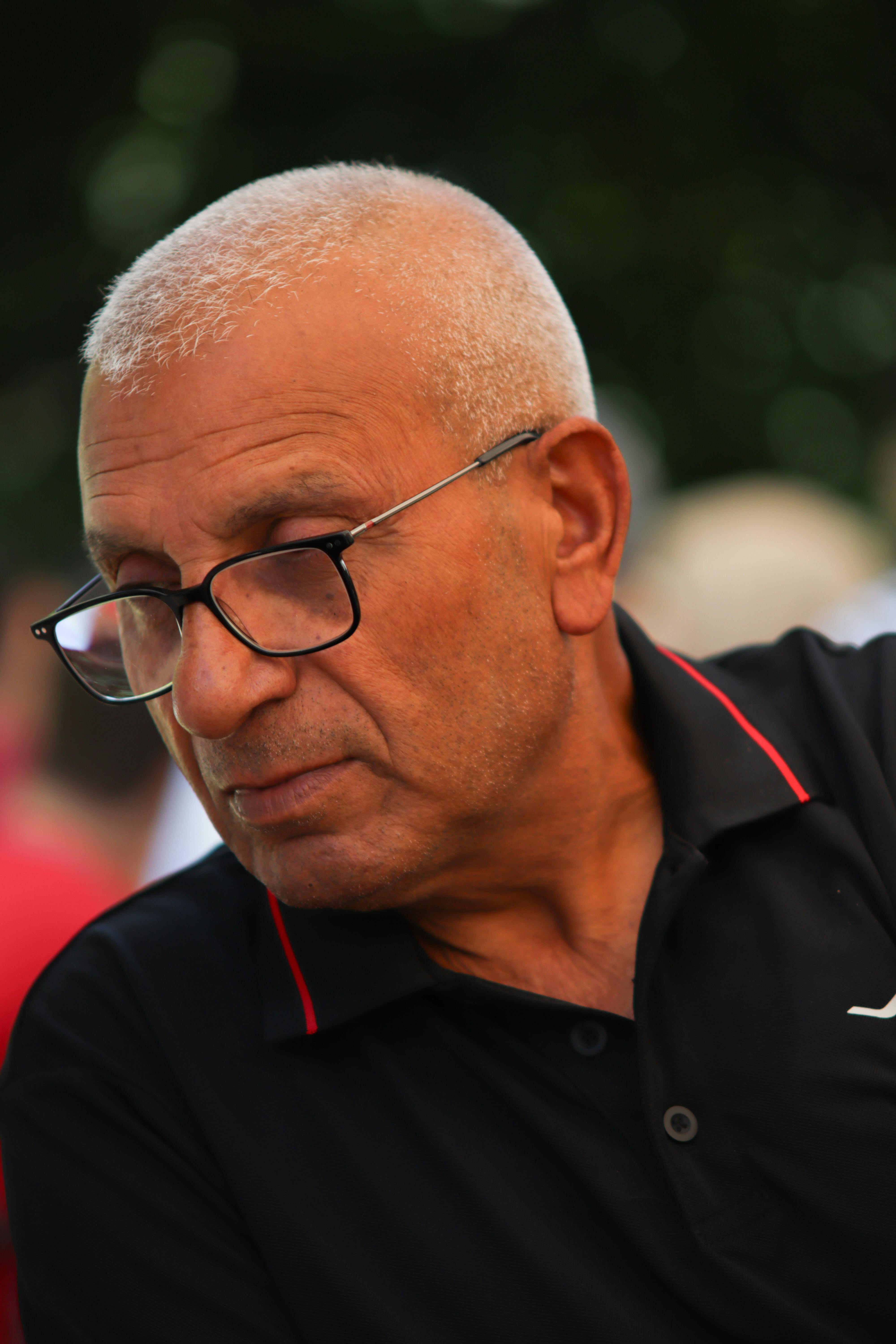 Elderly man with glasses in a thoughtful pose outdoors, wearing a black polo shirt.