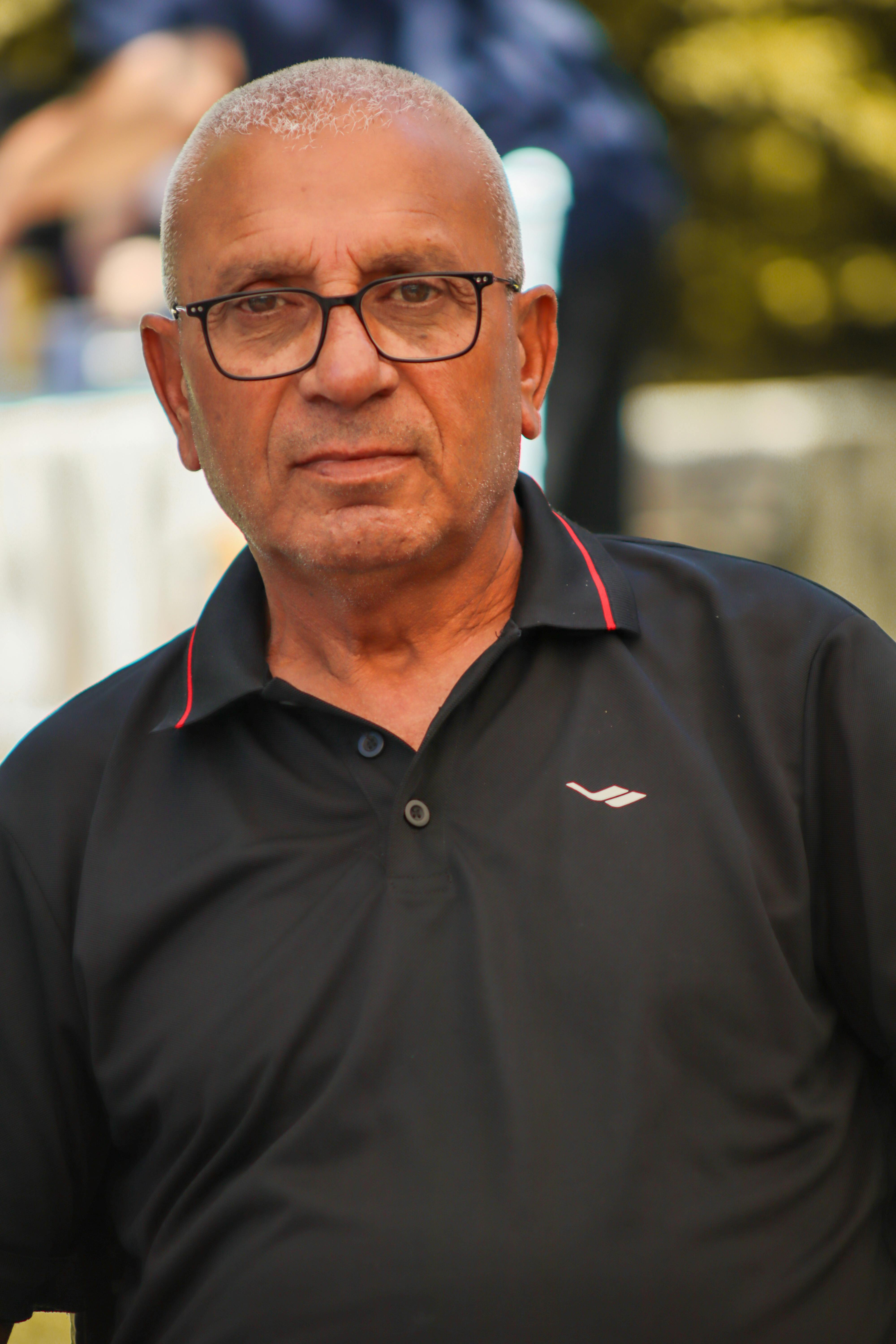 Portrait of an elderly man with eyeglasses in a black polo shirt, outdoors during the day.