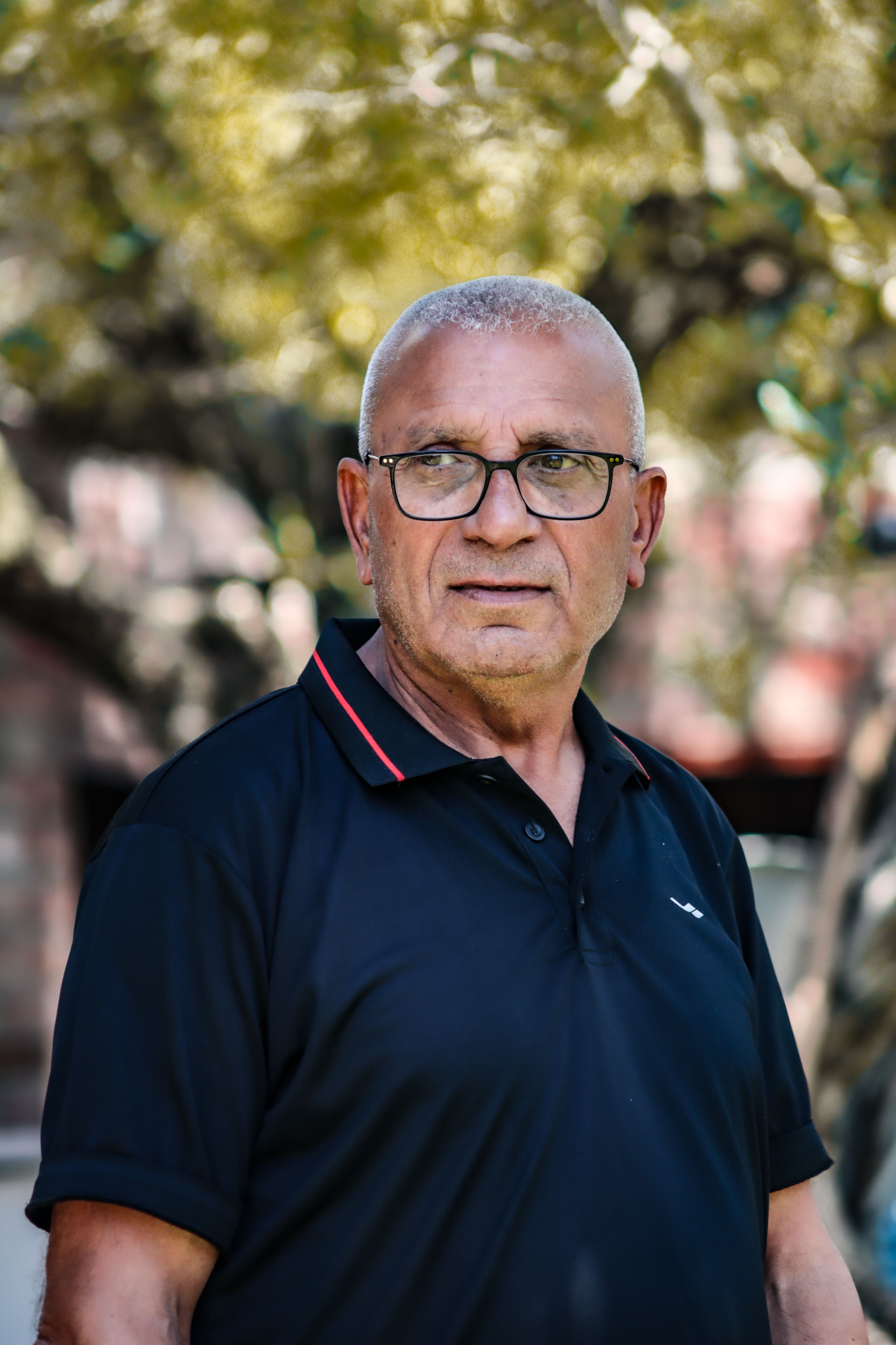 Portrait of an elderly man wearing glasses, standing in a park during the day.