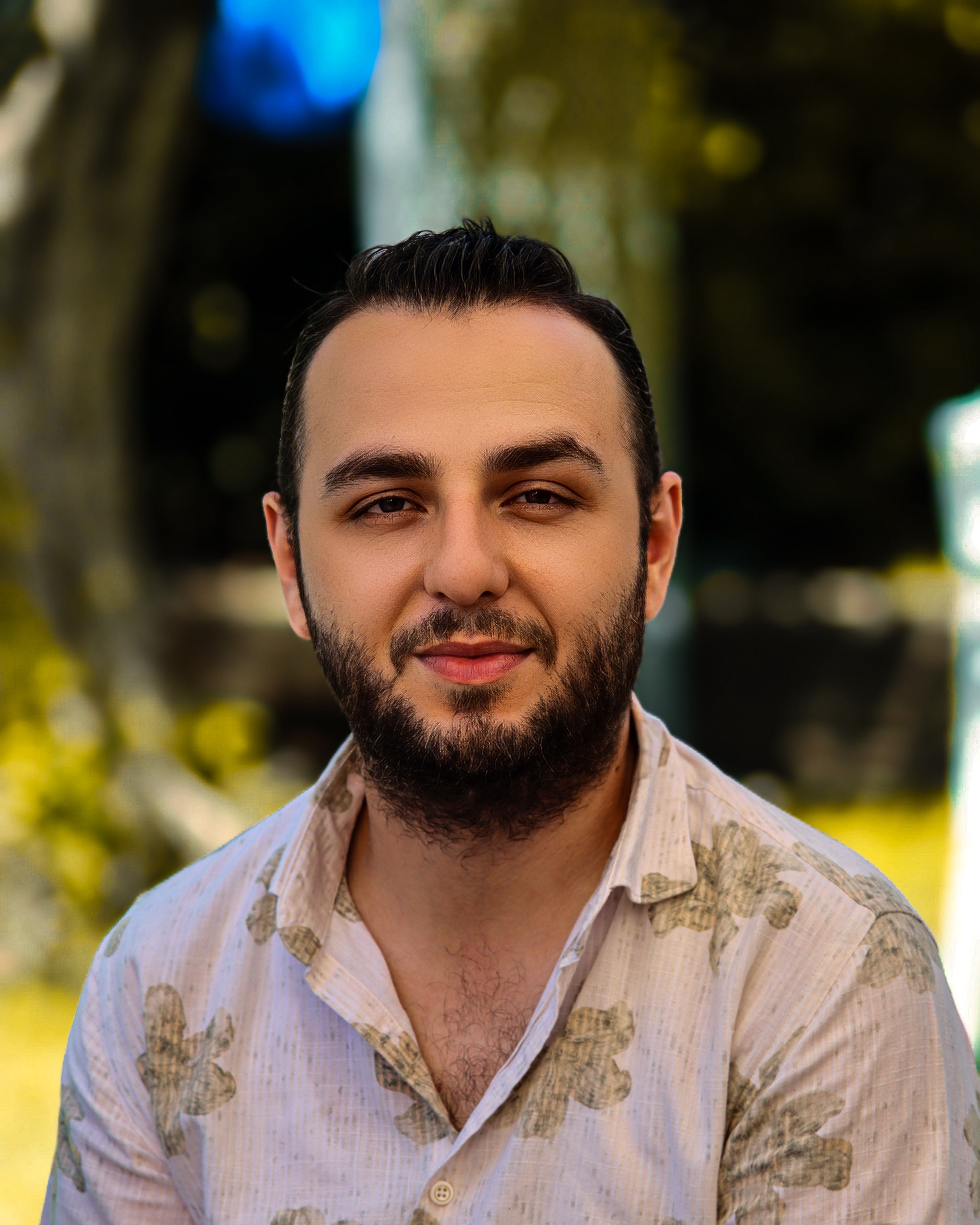A man with a beard smiles outdoors, surrounded by sunlight and trees.