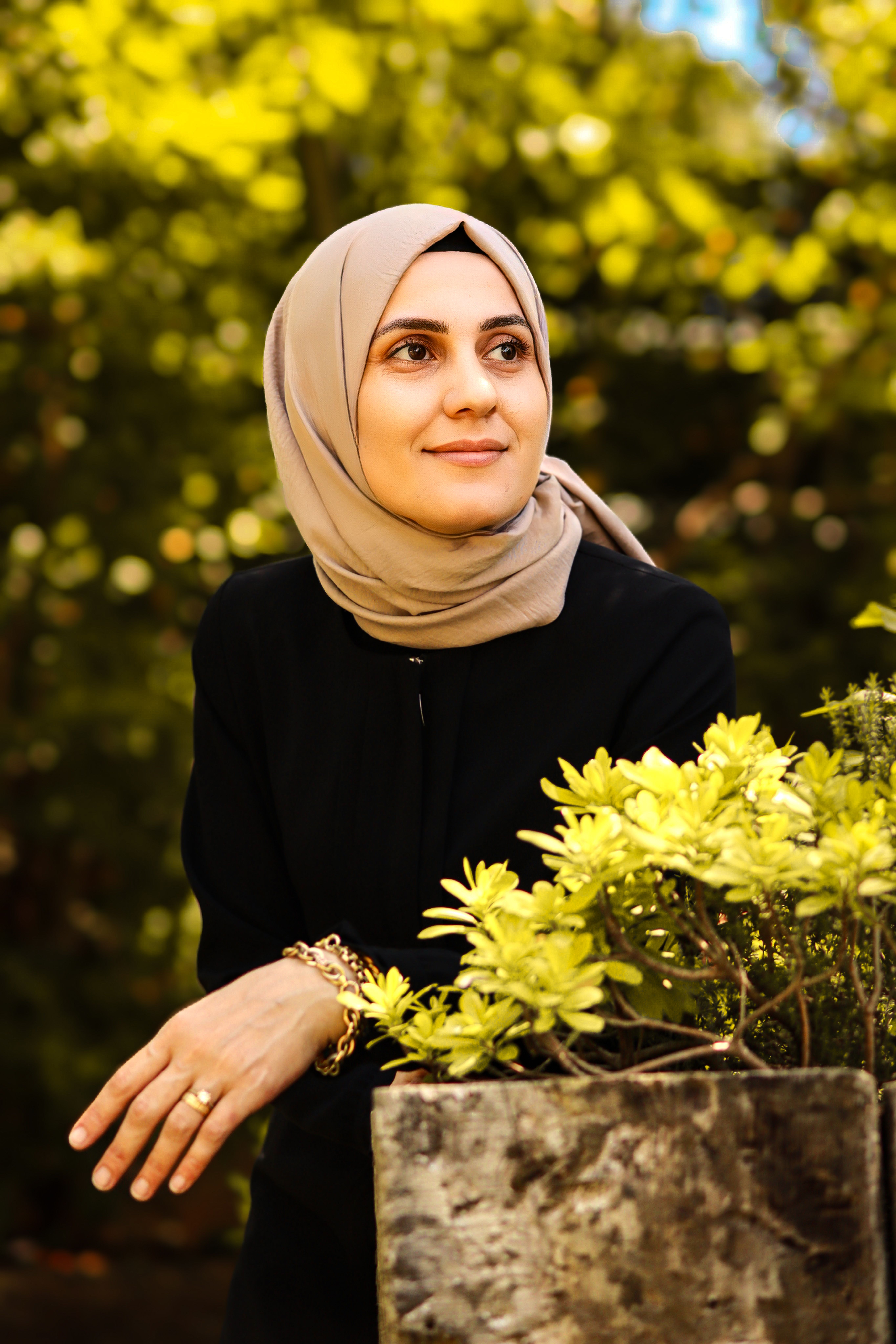 Elegant portrait of a woman in a beige headscarf surrounded by lush greenery in a park setting.