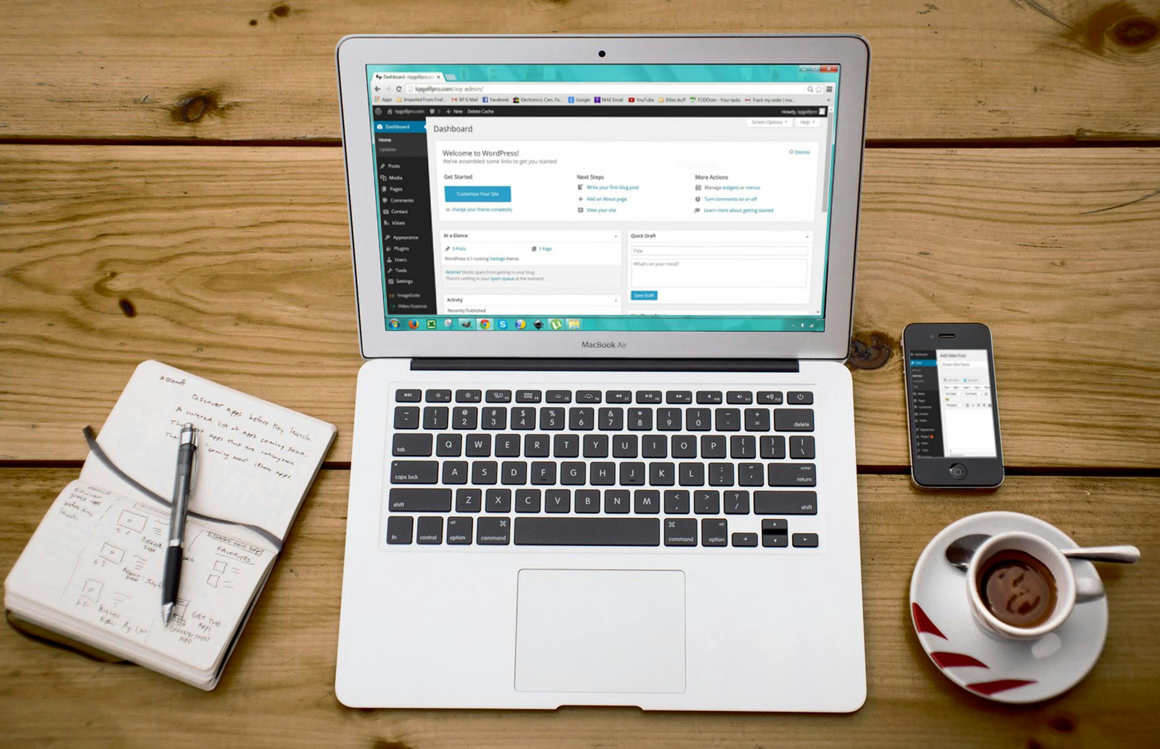 macbook screen displaying a wordpress dashboard, with a notebook on the left and a phone and a cup of coffee on the right