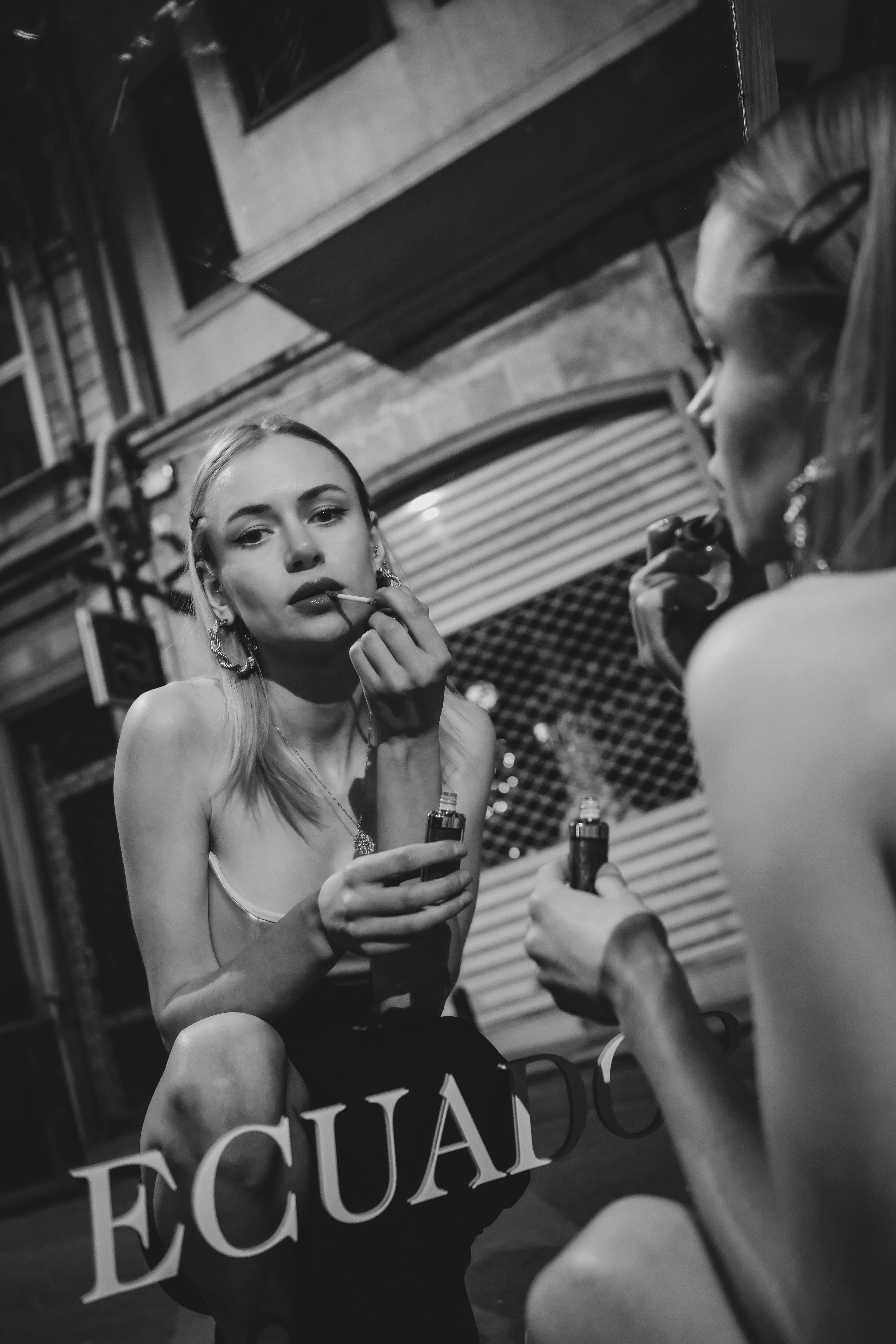 Black and white portrait of a woman applying makeup in city street reflection.