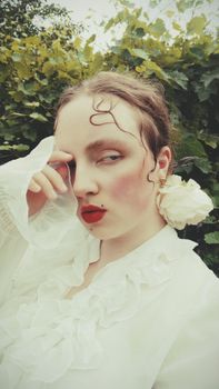 A serene portrait of a woman in a white ruffled blouse, adorned with a flower, set against lush greenery.