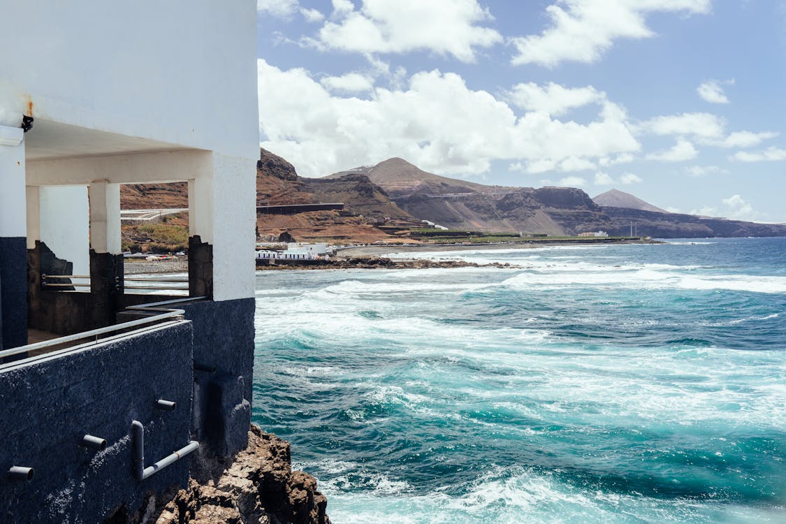 White and blue coastal structure overlooking turquoise waves with volcanic mountains and cloudy sky in the background.