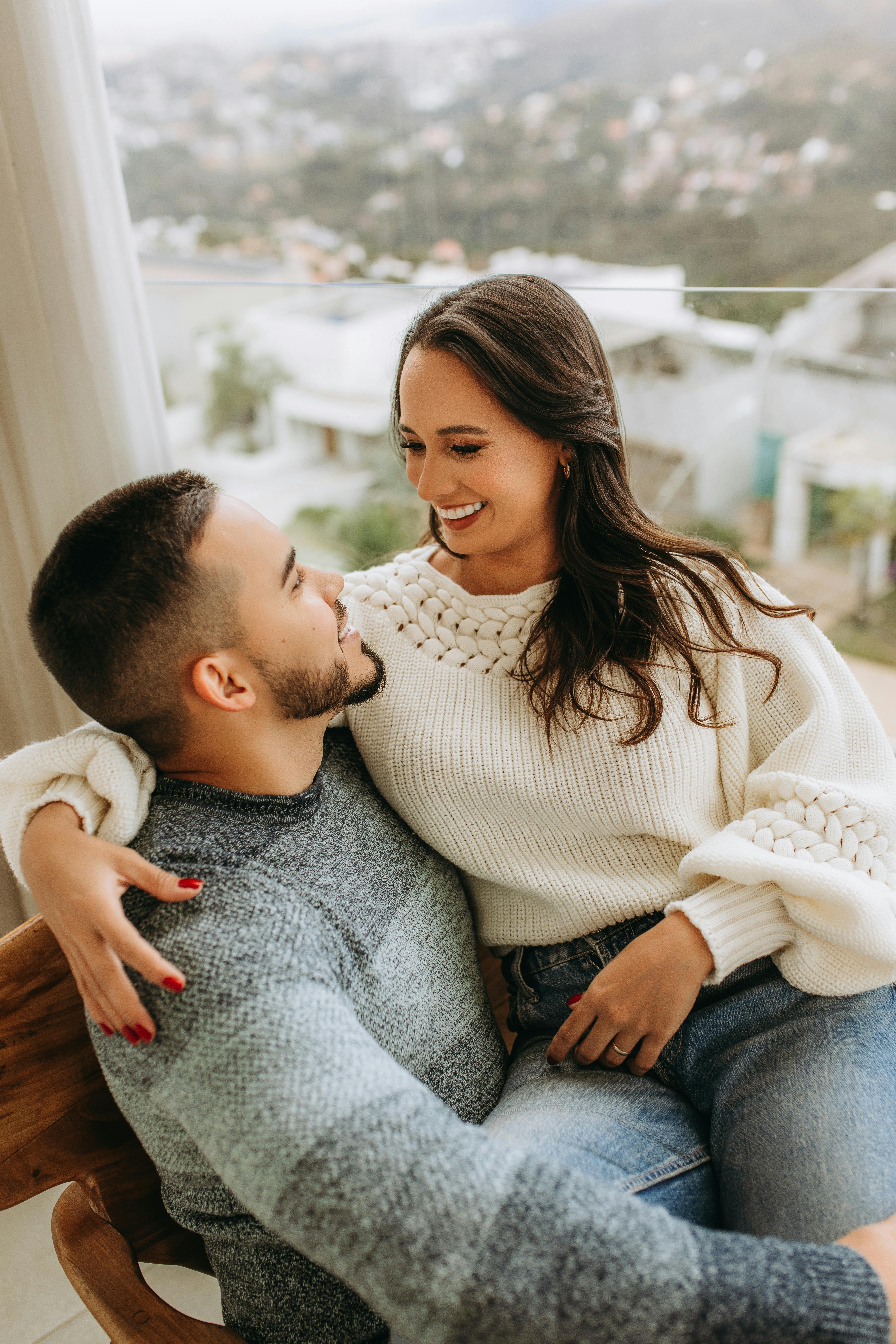 A loving couple in sweaters sharing a tender moment indoors with a view of Belo Horizonte.