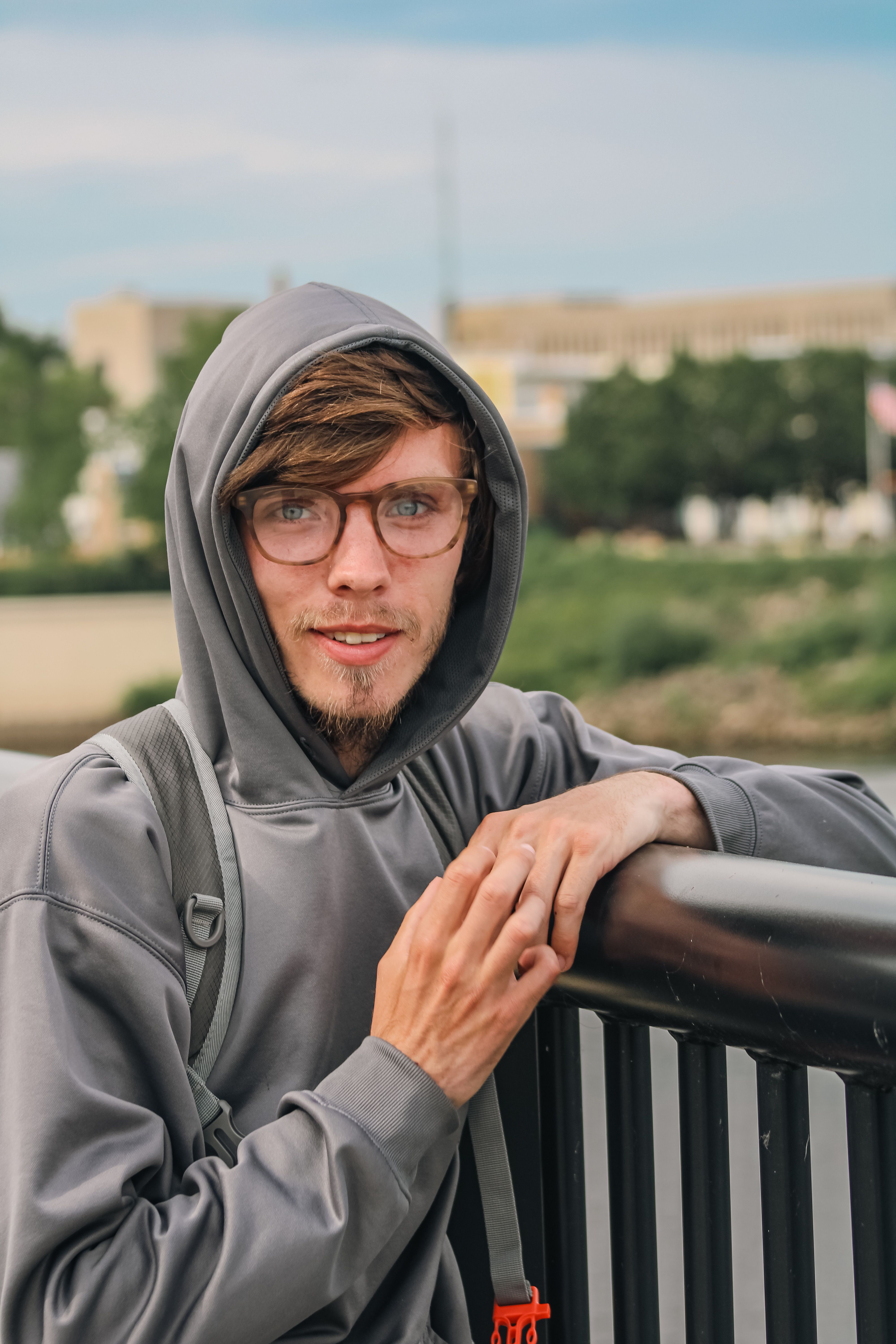 Young man in gray hoodie with glasses posing by outdoor railing, displaying a casual style.