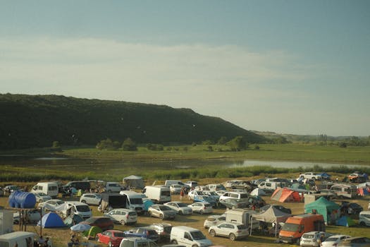 A lively camping festival with cars and tents by a scenic lake and mountains.