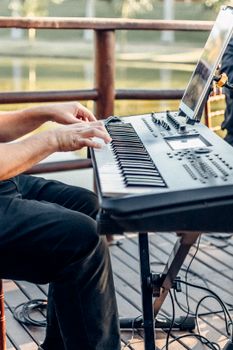 A musician plays an electronic keyboard with focus on hands, set against an outdoor backdrop.