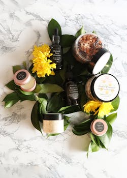 Top view of natural skincare products arranged with yellow flowers and greenery on marble surface.
