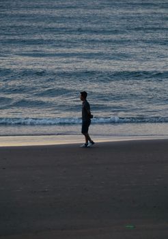 Silhouette of a man walking on a quiet beach during twilight with gentle waves in the background.