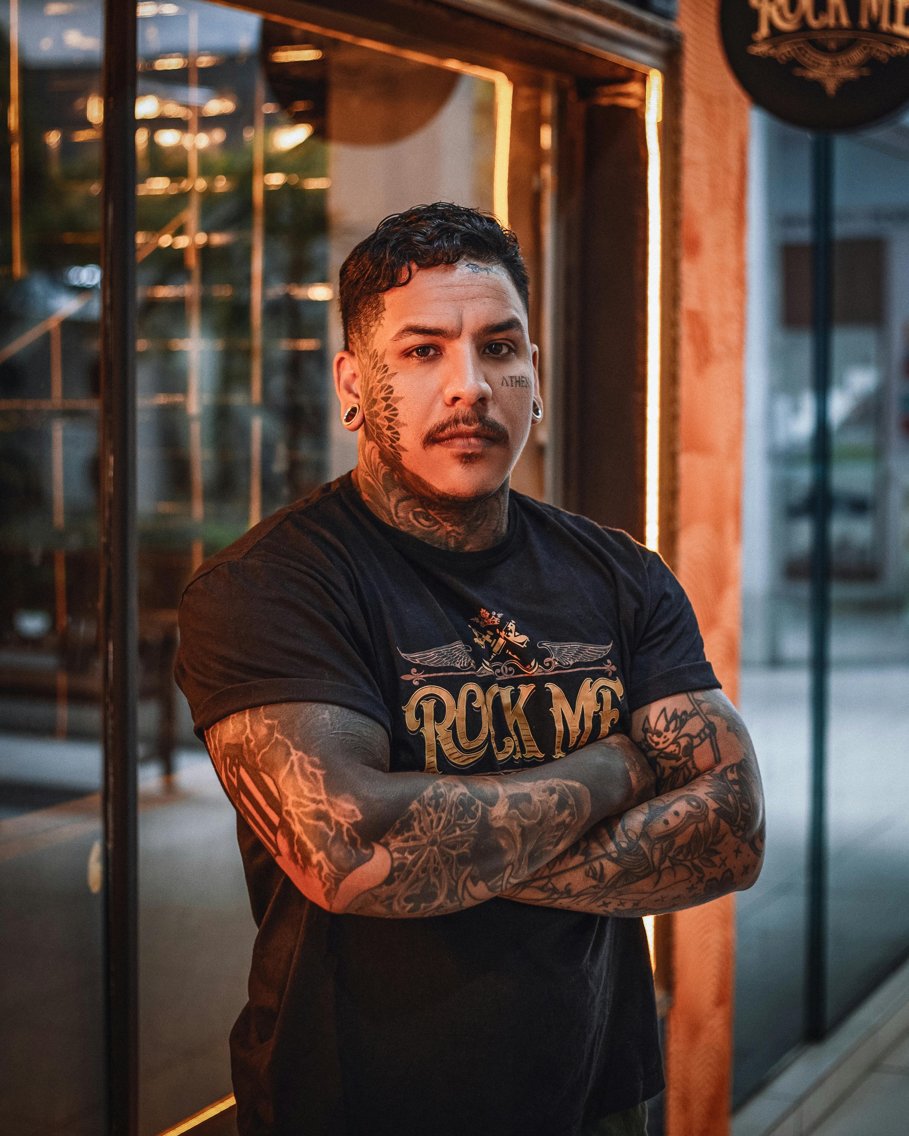 Portrait of a tattooed man in rock-themed fashion standing confidently with crossed arms in an urban setting.