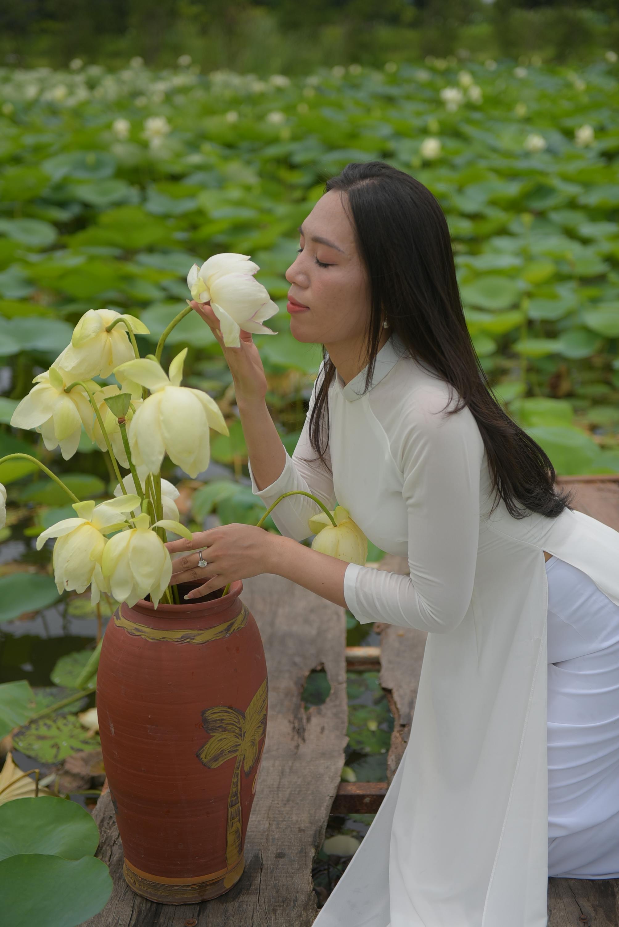 A graceful woman in a white Ao Dai kneels beside a vase of lotus flowers, embracing tradition and nature.