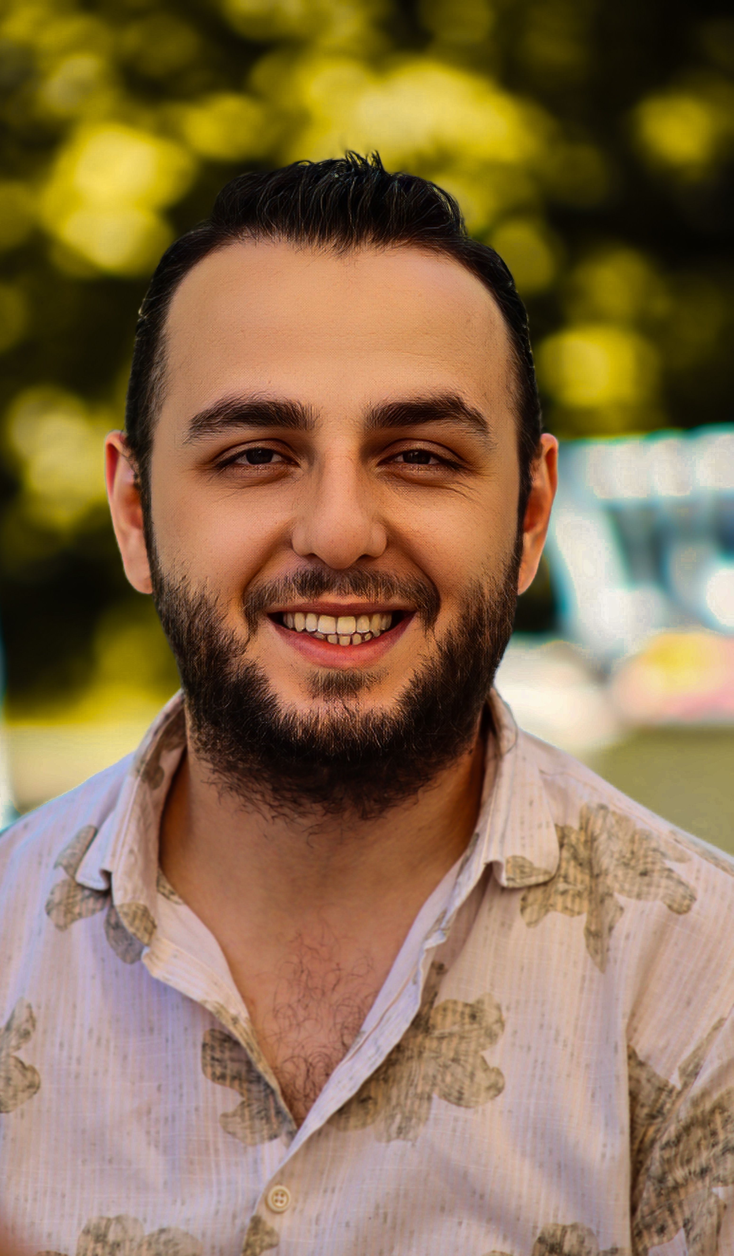 Cheerful man with a beard smiling outdoors in a sunlit park setting.