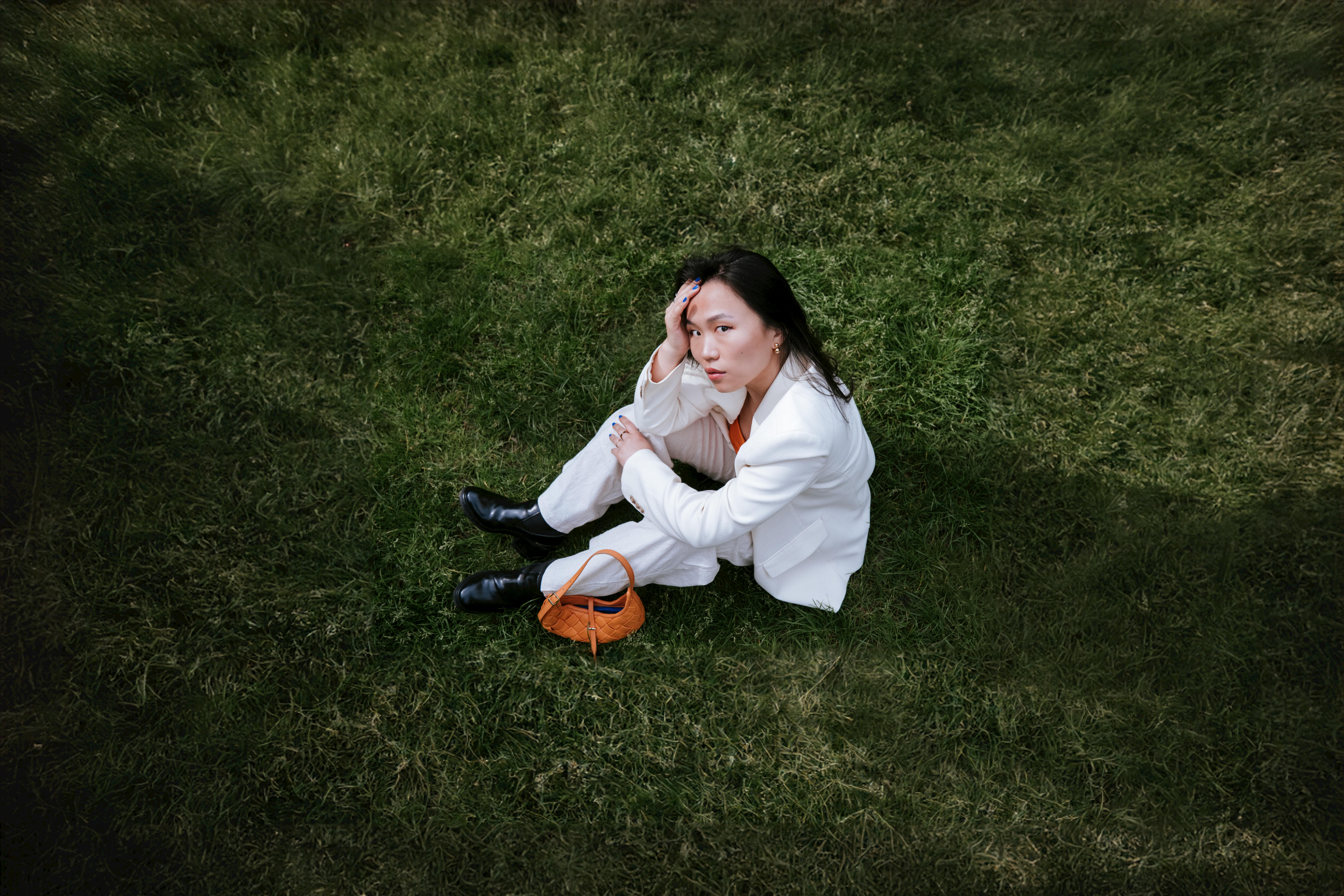 A stylish woman in a white jacket sits on the grass in Cologne, Germany, showcasing outdoor fashion.