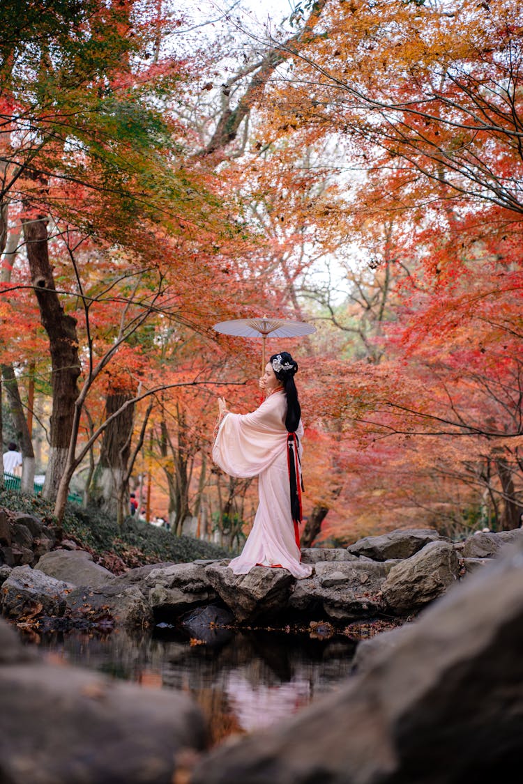 Woman In Traditional Clothing And With Umbrella Standing On Rocks Under Autumn Trees At Park