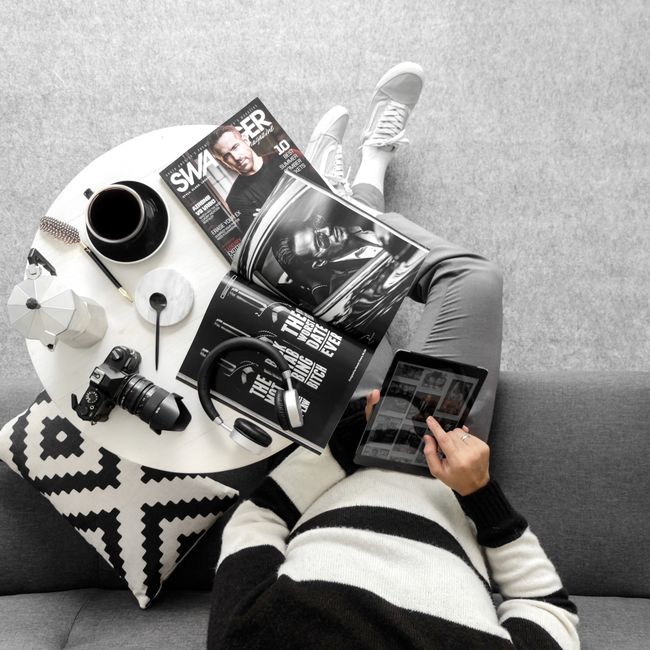 Man using a tablet while relaxing on a sofa with magazines and a camera on a side table.