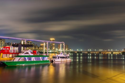 A serene night scene of illuminated city buildings and boats docked on a calm river.