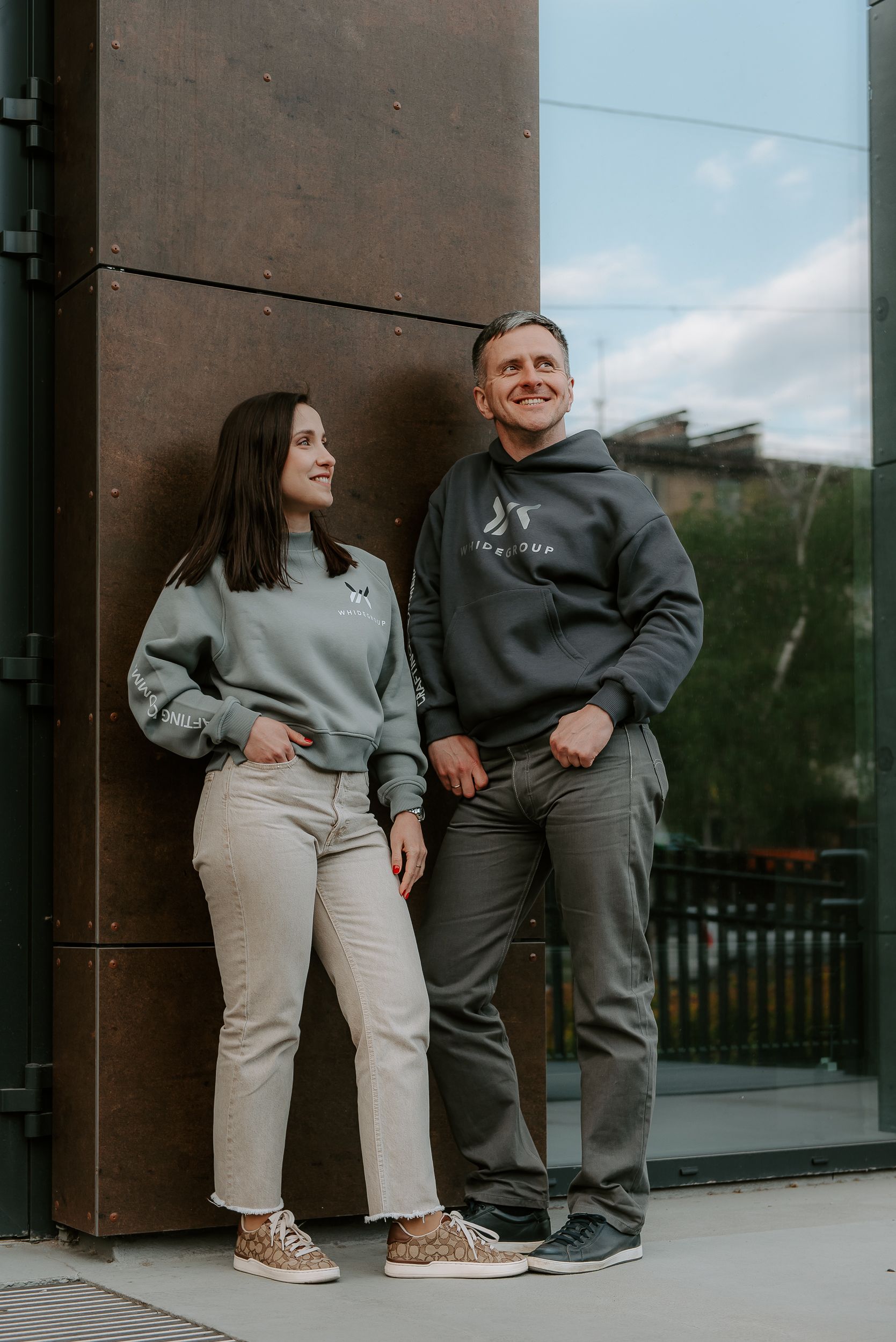 A happy couple wearing hoodies smiles in an urban outdoor setting, showcasing casual fashion.