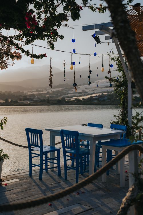 Free A serene outdoor dining setup by the river at sunset with blue chairs and hanging decorations. Stock Photo