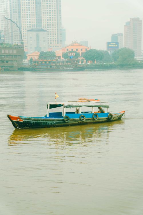 Free A scenic view of a traditional boat cruising on the Saigon River with Ho Chi Minh City's skyline. Stock Photo