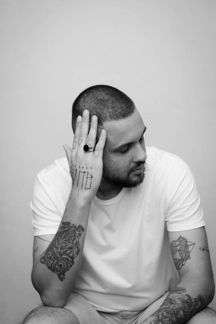 Black and white portrait of a tattooed man in a pensive pose indoors.