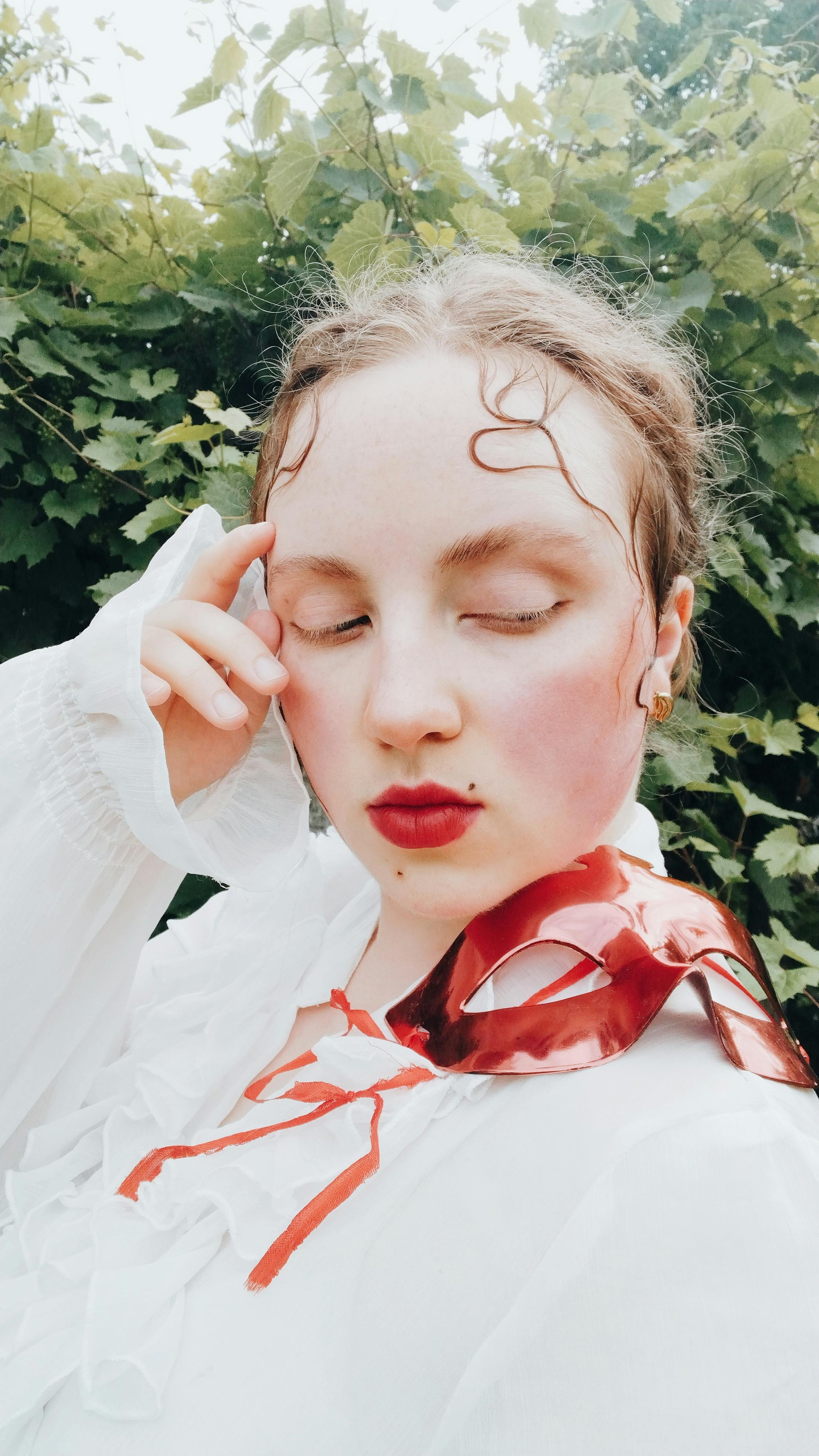 A striking portrait of a woman in a white blouse with a red mask, set against lush greenery.