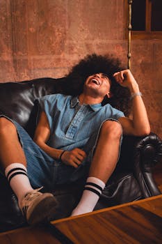 Model with afro hair in denim outfit laughing on a black sofa, showcasing fun and casual fashion.