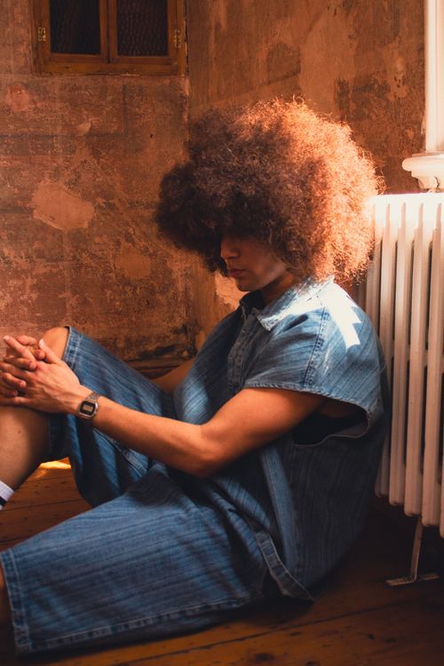 Free A contemplative man with afro-textured hair sits by a radiator in a rustic room, bathed in warm light. Stock Photo