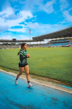 Brunette woman running on a blue track in a stadium, wearing athletic clothing under a clear sky.