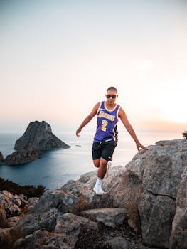 A man in a sports jersey explores rocky cliffs by the ocean at sunset, with a stunning coastal view.