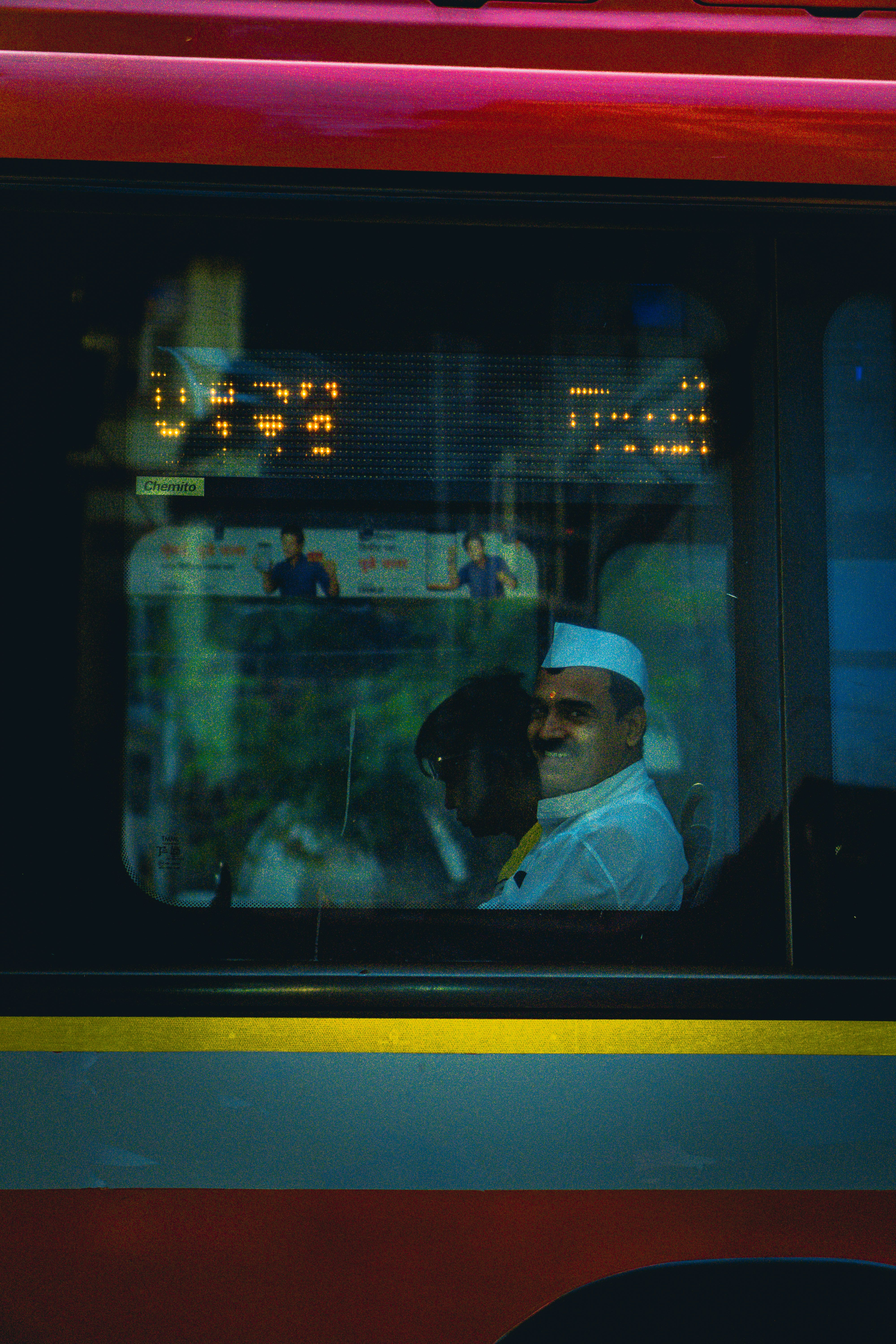 A man wearing a cap smiling while sitting inside a city bus visible through the window.