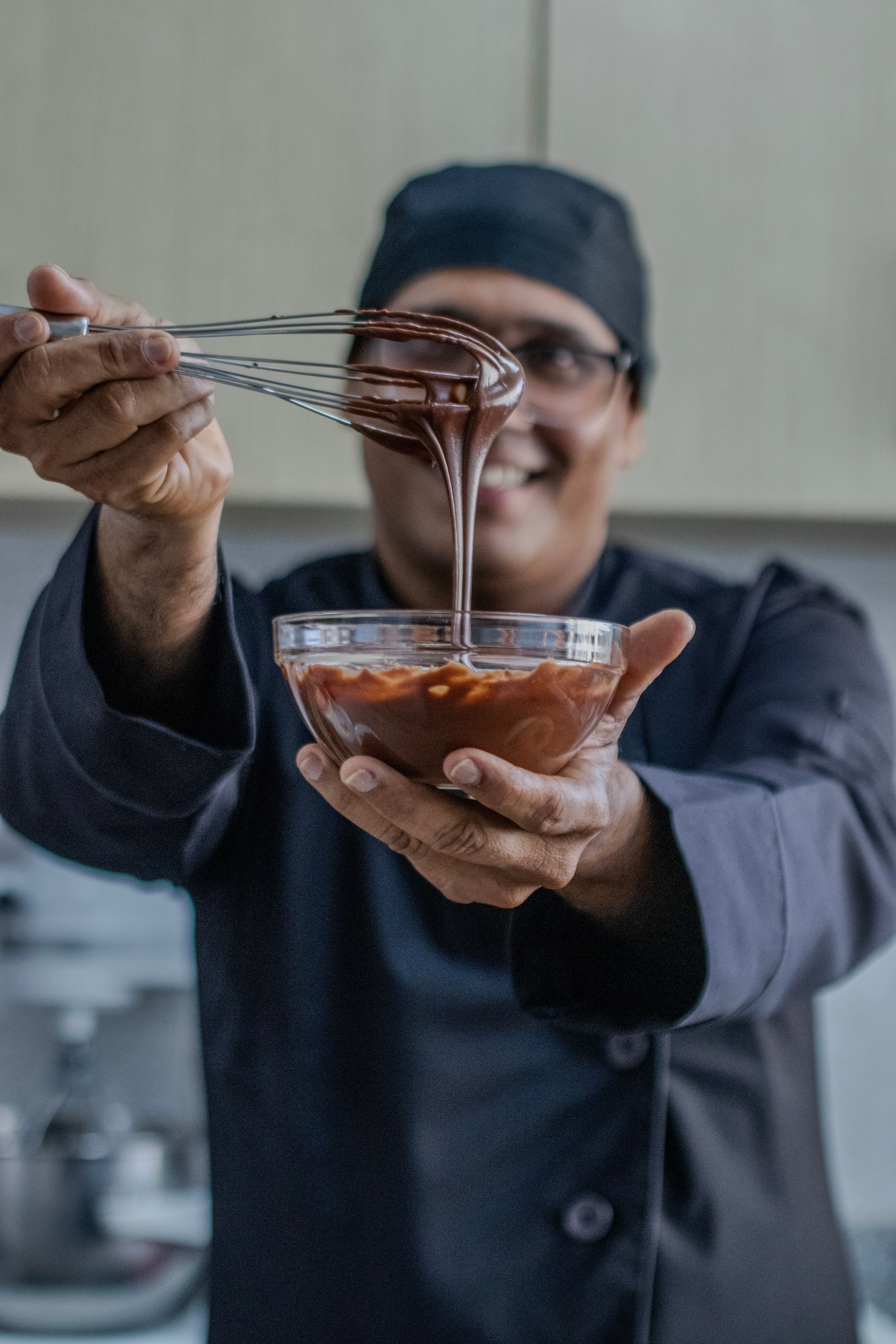 Chef smiling while holding a whisk and bowl with chocolate mixture in a kitchen.