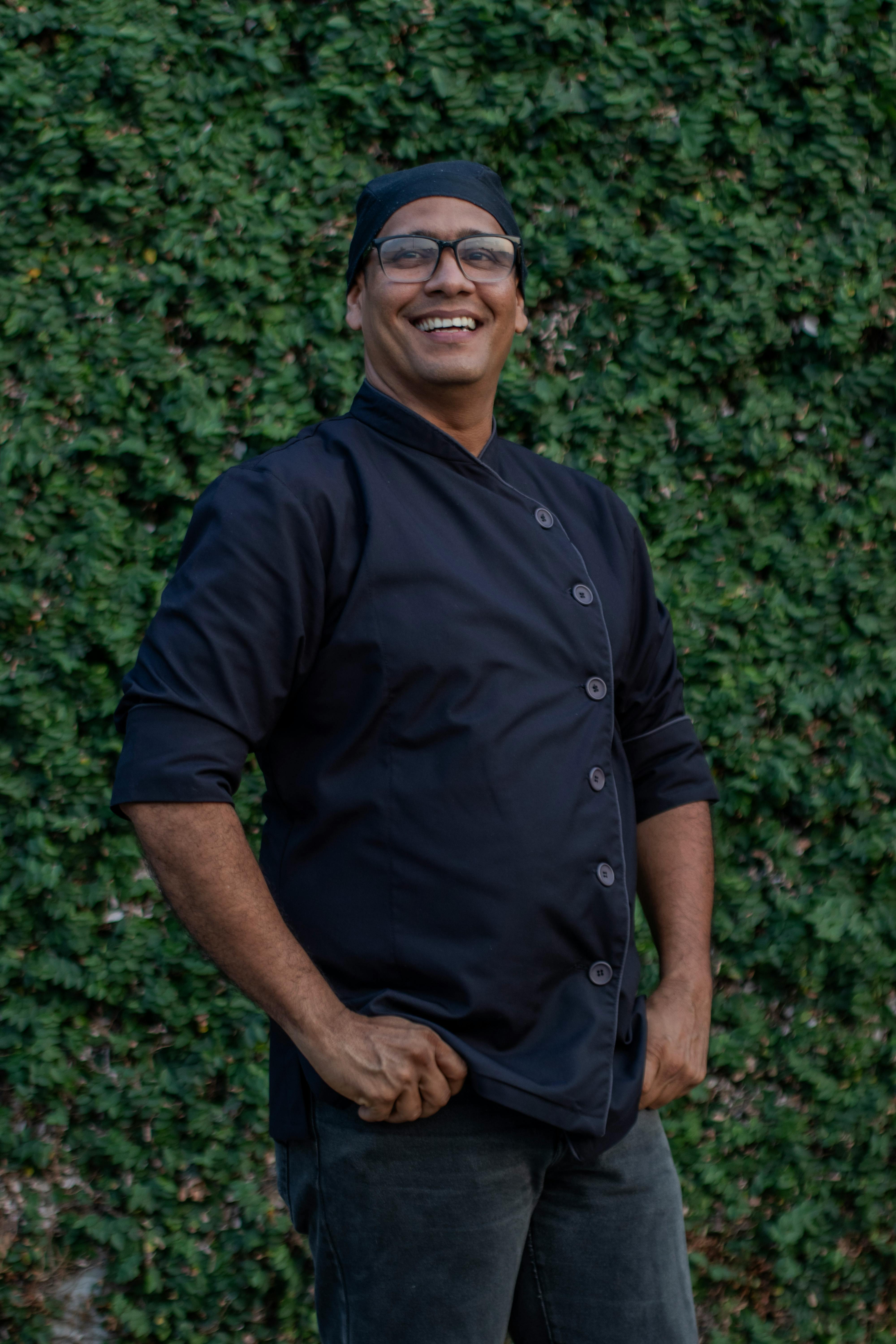 Smiling chef in black uniform standing outdoors against ivy wall.