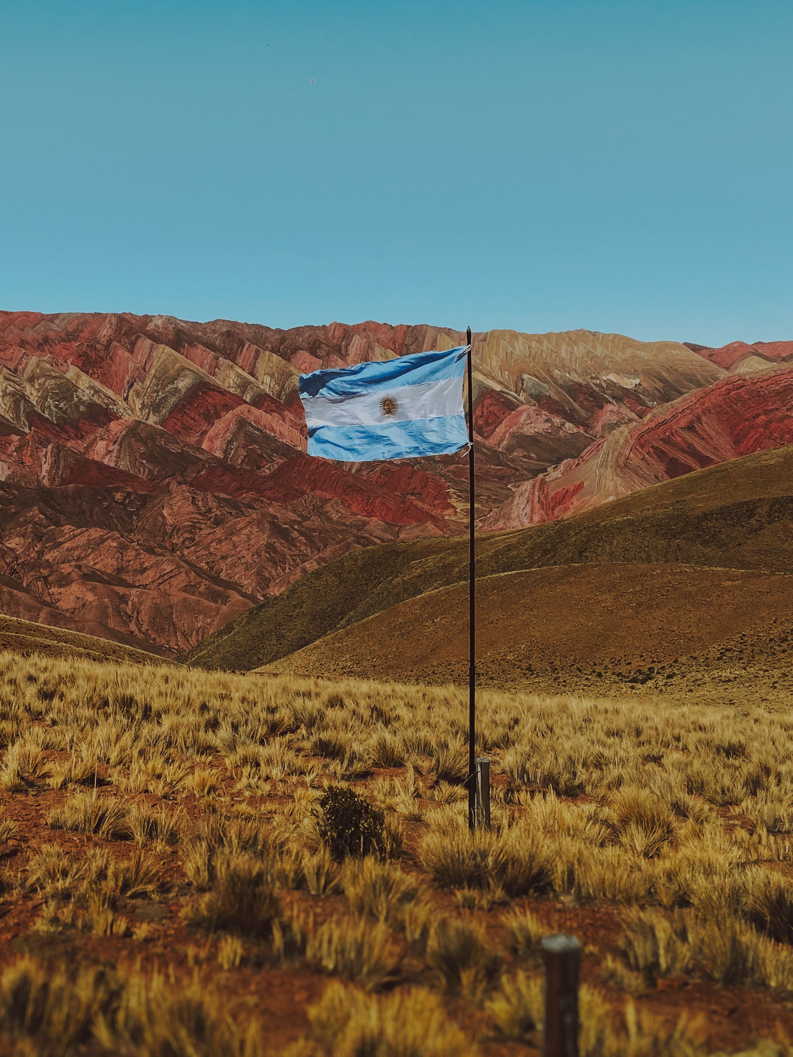 The Argentinian flag waving amidst the multicolored mountains and grassy plains of Argentina's rural countryside.