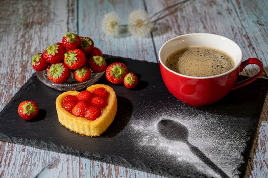 A delightful heart-shaped strawberry tart next to a cup of coffee on a rustic slate board.